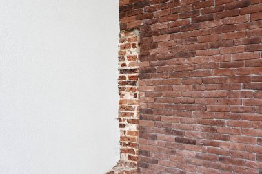 Beyaz alçı tabakasının arkasında kısmen korunmasız kalmış eski bir tuğla duvar. Kaba kırmızı tuğlalar ve pürüzsüz beyaz kaplama arasındaki tezat görsel ilgi ve doku ekler..