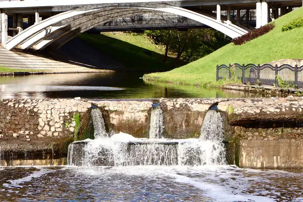 Taşın ve beton setin altında çağlayan küçük bir şelalesi olan şehir suyu. Arka planda, yayalar ve yol köprüleri yeşil bir parkta sakin bir nehri geçiyor..