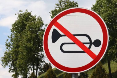 Kırmızı ile çizilmiş kornalı trafik işaretine yakın çekim. Yuvarlak tabela yeşil ağaçların ve parçalı bulutlu gökyüzünün arka planında duruyor..