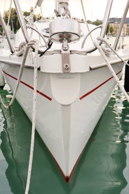 Yeşil suya kenetlenmiş kırmızı çizgili beyaz bir yelkenlinin önünde. Halatlar ve metal parçaları teknelerin temiz tasarımı ve denizcilik detaylarını vurguluyor..