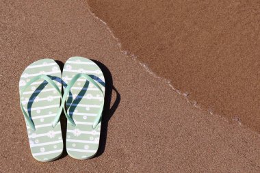 Yumuşak bir dalga yaklaşırken, kumlu sahilde papatya desenli açık yeşil bir çift parmak arası terlik duruyor. Fotoğraf rahatlatıcı bir yaz gününün özünü yakalıyor..