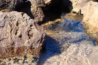 Görüntü, kıyı kayalarının kristal berrak suya kısmen battığını gösteriyor. Güneş ışığı dalgalanan yüzeyden yansıyarak aşağıdaki deniz tabanını ve deniz dokusunu ortaya çıkarır..