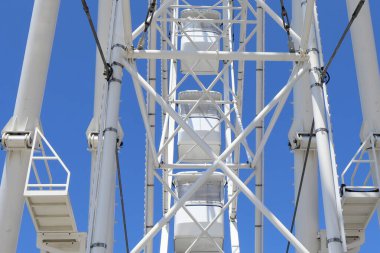 Geometrik metal destekleri ve kulübeleri olan beyaz dönme dolap yapısının alçak açılı görüntüsü canlı mavi gökyüzüne karşı kurulmuş. Görüntü, mimari simetriyi ve yaz tatilini yansıtıyor.. 