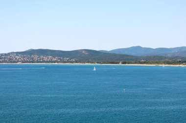 Sakin mavi sularda yüzen küçük bir yelkenlinin olduğu huzurlu bir sahil manzarası. Uzak kıyı şeridi yeşil tepelerle kaplı ve gökyüzünün altında dağınık evler..