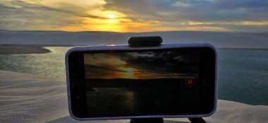 Esnek bir tripoda monte edilmiş akıllı bir telefon çöl kumunun el değmemiş beyaz kumlarında oturur ve sakin bir su kütlesine yansıyan çarpıcı bir günbatımını yakalar. Gökyüzü yumuşak pastel renklerle boyanmıştır..