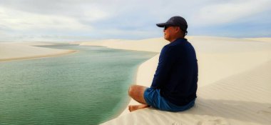 Beyaz kum tepecikleri çölündeki bir göl kenarında rahatlayan bir adam. Mavi gökyüzü kumla güzel bir kontrast oluştururken, güneş kristal berrak sudan yansıyarak yumuşak bir şekilde parlar. Sahne, sakinliği ve doğayla bağlantıyı iletir..