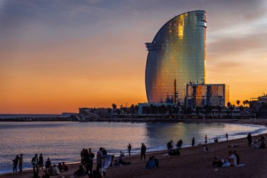 Barselona, İspanya-19 Şubat 2024. Barceloneta sahilinde dinlenen insanlar, canlı bir günbatımında, ikonik w Barselona oteli gökyüzünün sıcak renklerini yansıtıyor.