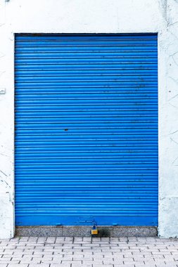 Altında asma kilit bulunan yatay çizgili, kapalı mavi bir kepenk kapağı. Kapı beyaz bir duvara dayanıyor, bir tarafında desenli bir yüzey var..