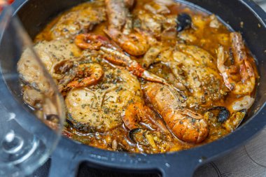 Siyah bir tencerede deniz ürünleri güvecinin yakın plan çekimi, zengin bir et suyunda karides, balık ve otlar var..