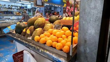 Canlı bir market tezgahında taze portakal ve kavunlar sergilenirken, arka planda müşteriler geziniyor. Meşgul bir pazarda tipik bir meyve standı.