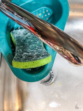 Paslanmaz çelik mutfak lavabosu, sabunlu su, yeşil sünger ve musluğun altındaki mavi drenaj sepetinin yakın çekimi..