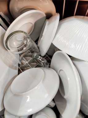 Temiz beyaz tabaklar, çay tabakları ve bulaşık makinesinin içine istiflenmiş cam bardakların yakın plan görüntüsü, çeşitli bulaşık takımları şekilleri ve dokuları düzenli bir düzende sergiliyor..