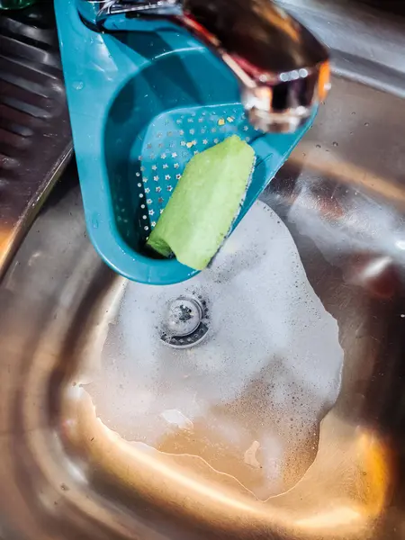 Paslanmaz çelik mutfak lavabosu, sabunlu su, yeşil sünger ve musluğun altındaki mavi drenaj sepetinin yakın çekimi..