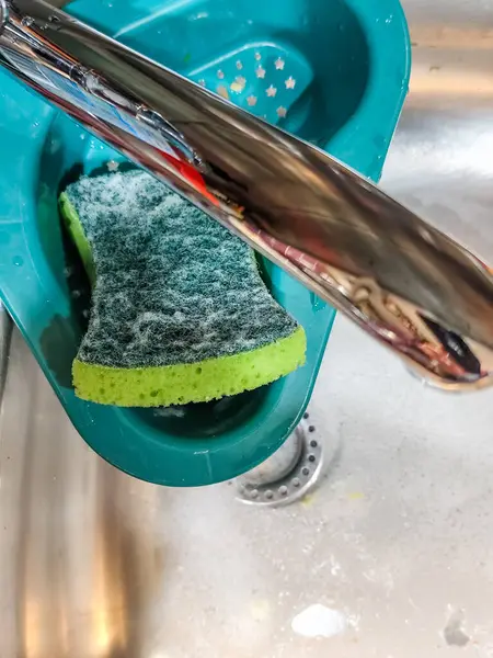 Paslanmaz çelik mutfak lavabosu, sabunlu su, yeşil sünger ve musluğun altındaki mavi drenaj sepetinin yakın çekimi..