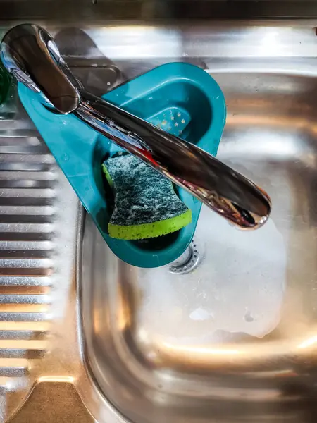 Paslanmaz çelik mutfak lavabosu, sabunlu su, yeşil sünger ve musluğun altındaki mavi drenaj sepetinin yakın çekimi..