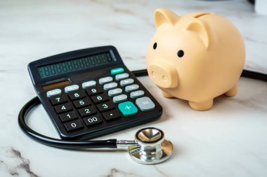 Hesap makinesi ve steteskobu olan bir kumbara sağlık hizmetlerinin finansal yönünü gösteriyor..