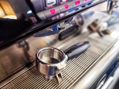 Ticari bir espresso makinesinin, metal filtre sepetine ve sapına odaklı bir iskele filtresinin yakın çekimi. Arka planda düğmeleri olan makinenin kontrol paneli görünür.