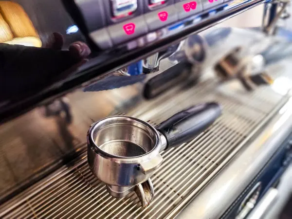 Ticari bir espresso makinesinin, metal filtre sepetine ve sapına odaklı bir iskele filtresinin yakın çekimi. Arka planda düğmeleri olan makinenin kontrol paneli görünür.