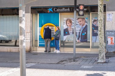 Barselona, İspanya-4 Haziran 2024. Müşteriler güneşli bir şehirde CaixaBank ATM 'sinde para transferi yapıyor..