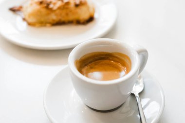 Tabakta beyaz bir fincan espresso, arka planda bulanık kremalı pasta, hayatın basit bir zevkini yakalıyor.