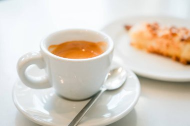 Tabakta beyaz bir fincan espresso, arka planda bulanık kremalı pasta, hayatın basit bir zevkini yakalıyor.