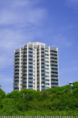 Beyaz bir meskun gökdelen, açık mavi gökyüzüne karşı araziye hükmediyor, aşağıdaki yeşil şehir parkına tamamen zıtlık sunuyor.