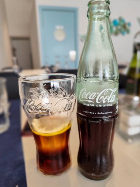 Barselona, İspanya-12 Mayıs 2025. Klasik bir Coca-Cola şişesinin yanında buzla dolu bir bardak Coca-Cola. tablo ve bulanık arkaplan ögeleri.