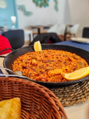 Limon dilimleriyle süslenmiş bir tava deniz mahsulü paella morina balığı. Restoran ortamında örülmüş bir paspasın üzerinde servis ediliyor..