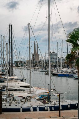 Badalona, İspanya-1 Ocak 2025. Badalona Limanı 'na yanaşan yelkenli ve yatların arkasında endüstriyel bacalar ve şehir binaları bulutlu gökyüzünün altında..