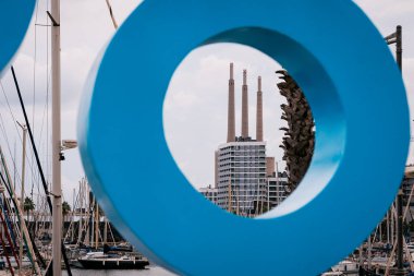 Badalona, İspanya-1 Ocak 2025. İkonik Badalona bacalarının mavi dairesel bir heykelle çerçevelenmiş ön planda yelkenli ve marina manzarası.