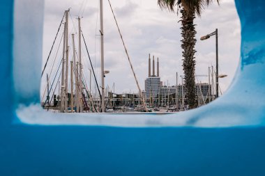 Badalona, İspanya-1 Ocak 2025. İkonik Badalona bacalarının mavi dairesel bir heykelle çerçevelenmiş ön planda yelkenli ve marina manzarası.