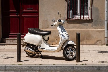 Klasik beyaz scooter, kırmızı kapılı ve parmaklıklı pencereli eski bir Avrupa binasının önündeki güneşli şehir kaldırımına park edilmiş..