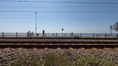 Badalona, İspanya-25 Ağustos 2025. İnsanlar Badalona sahilinde, İspanya 'da tren rayları, palmiye ağaçları, Akdeniz ve mavi gökyüzü olan yaz tatilinin tadını çıkarıyorlar. Zaman aşımı