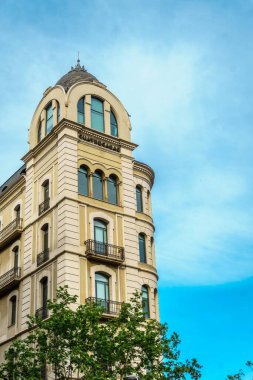 Göz kamaştırıcı bir modernist bina parlak mavi Barselona gökyüzüne karşı duruyor, yemyeşil bir ağaç ve tüylü beyaz bulutlar tarafından çerçevelenmiş..