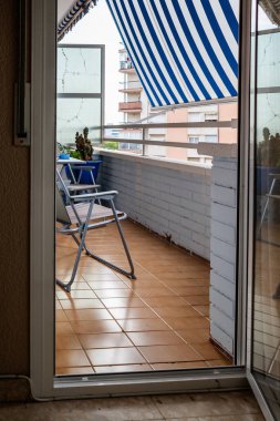 A small balcony with a folding chair, potted plants, and a striped awning.