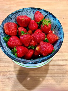 Benekli mavi bir seramik kasede taze kırmızı çilek manzarası. Kase beyaz bir tabakta duruyor. Tahta bir yüzey üzerinde yeşil bir halka var..