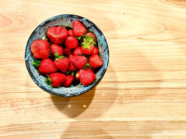 Bir kase olgun kırmızı çilek açık ahşap bir yüzeye oturur. Mavi seramik kase, doğal ahşap tanecikleri ve canlı meyvelerle çelişiyor..