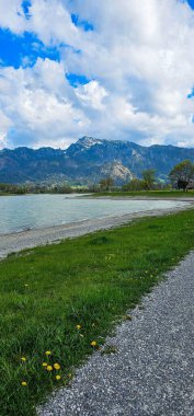 Görkemli dağlarla çevrili sakin bir göl manzarası. Gökyüzü parçalı bulutlu, mavi gökyüzüne karşı beyaz bulutlar, yeşil çimenler ve önplandaki sarı çiçekler parlak renkli patlamalar eklerken,.