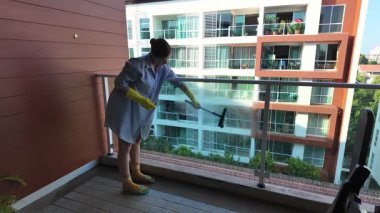 Mavi gömlekli ve sarı eldivenli kadın balkon camını temizliyor.