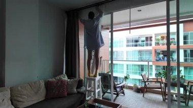 Mavi gömlekli ve sarı eldivenli kadın balkon camını temizliyor.