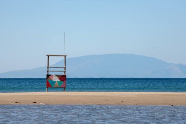 Renkli geometrik çizimlere sahip boş bir cankurtaran kulesi, sezon dışında Yunanistan 'da sakin bir plajda duruyor. Sakin deniz ve uzak dağlar sakin, minimalist bir kıyı manzarası yaratır..