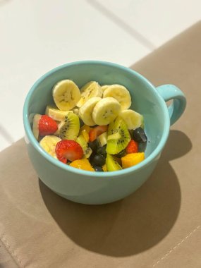 Muz, çilek, kivi ve mango gibi lezzetli taze meyve karışımlarıyla dolu mavi bir kupa canlı renkler ve sağlıklı tatlar yaratıyor..