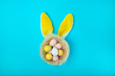 Yaratıcı bir Paskalya yuvası renkli dekoratif yumurtalarla dolu, canlı mavi arka planda neşeli tavşan kulaklarıyla şekillendirilmiş. Şenlikli ve eğlenceli bir tatil konsepti Mevsimlik tasarımlar için mükemmel.