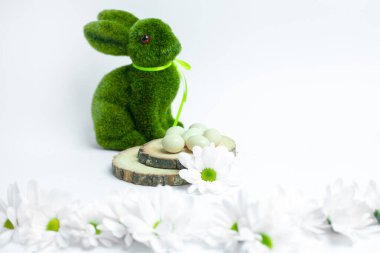 Beyaz arka planda beyaz papatyalarla çevrili beyaz çikolata şekerli yumurtaların arkasında kurdeleli yeşil yosun tavşanı. Mevsimsel promosyonlar ve içerik için mükemmel Paskalya ya ya da bahar dekoru sahnesi.