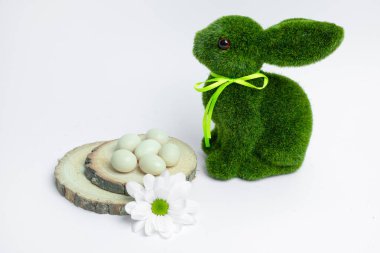 Pastel şeker yumurtalarının yanında kurdeleli yeşil yosun tavşanı ve tahta dilimlerinde papatya çiçeği. Bahar dekorasyonu, festival promosyonları veya mevsimsel içerik için minimum Paskalya kompozisyonu.