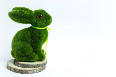 Kurdeleli yeşil yosun kaplı tavşan heykelciği, beyaz arka planda tahta bir kesiğin üzerinde oturuyor. Paskalya, bahar dekorasyonu, doğa temalı iç mekanlar ve şenlikli mevsimlik gösteriler için ideal..