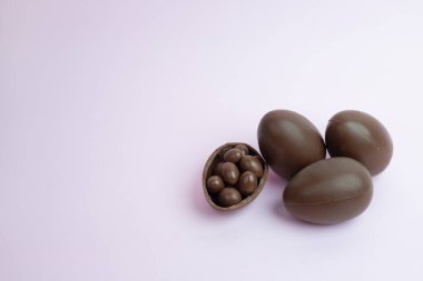 Yumuşak pastel arka planda dört çikolatalı Paskalya yumurtası ve içinde çikolata kaplı şekerler olan kırık bir kutu. Mevsimsel promosyonlar ve tatlı ikramlar için lezzetli ve şenlikli bir imaj..