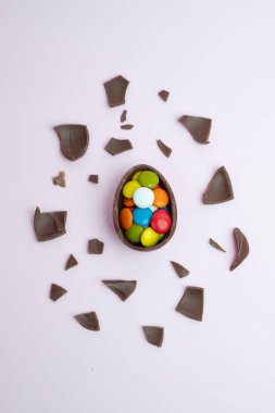 Çatlamış çikolata yumurtasının yukarıdan çekimleri canlı, renkli şekerlemelerle dolu, pastel pembe arka planda çikolata parçalarıyla çevrili, bayram kutlamaları için mükemmel..