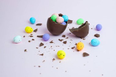 Çatlak bir çikolata yumurtası rengarenk benekli mini yumurtaları dağınık çikolata parçalarıyla çevrili pastel bir yüzeye döküyor. Bayramlık Paskalya temalı bir ikram konsepti.
