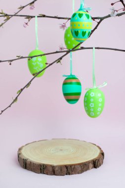 Karmaşık desenli yeşil ve sarı Paskalya yumurtaları ağaç dalından sarkan ahşap bir platformun üzerinde. Yenilenmeyi ve geleneği sembolize eden doğal bir bayram düzenlemesi..